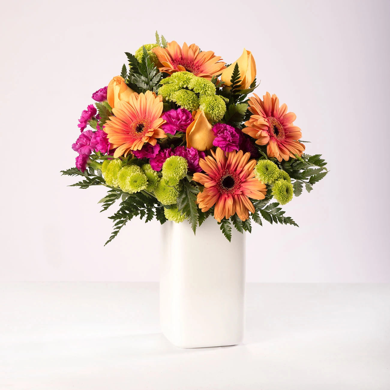 Arreglo de gerberas y rosas anaranjadas, acompañado de claveles fucsias y flor pinocho, en un florero.