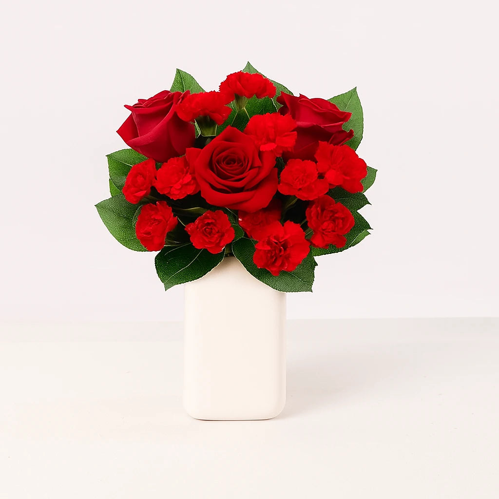 Arreglo floral con rosas y claveles rojos en florero blanco o de vidrio transparente.