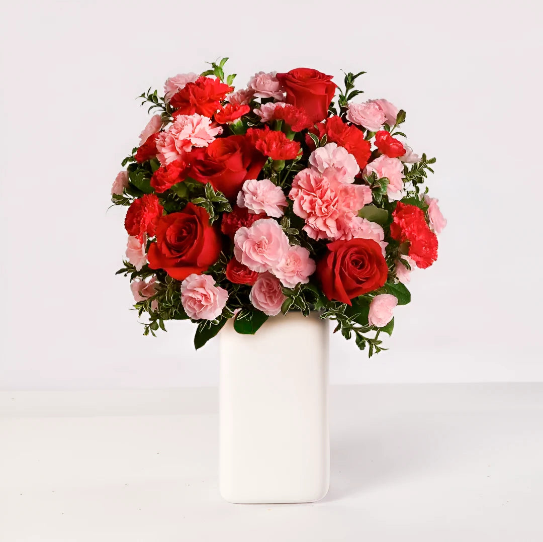 Arreglo floral con rosas rojas, claveles rojos y rosados, con follaje verde sobre un florero.