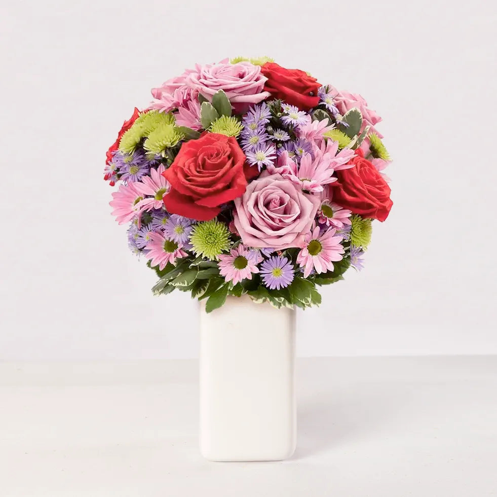 Arreglo de rosas rojas y rosadas con margaritas y follaje verde.